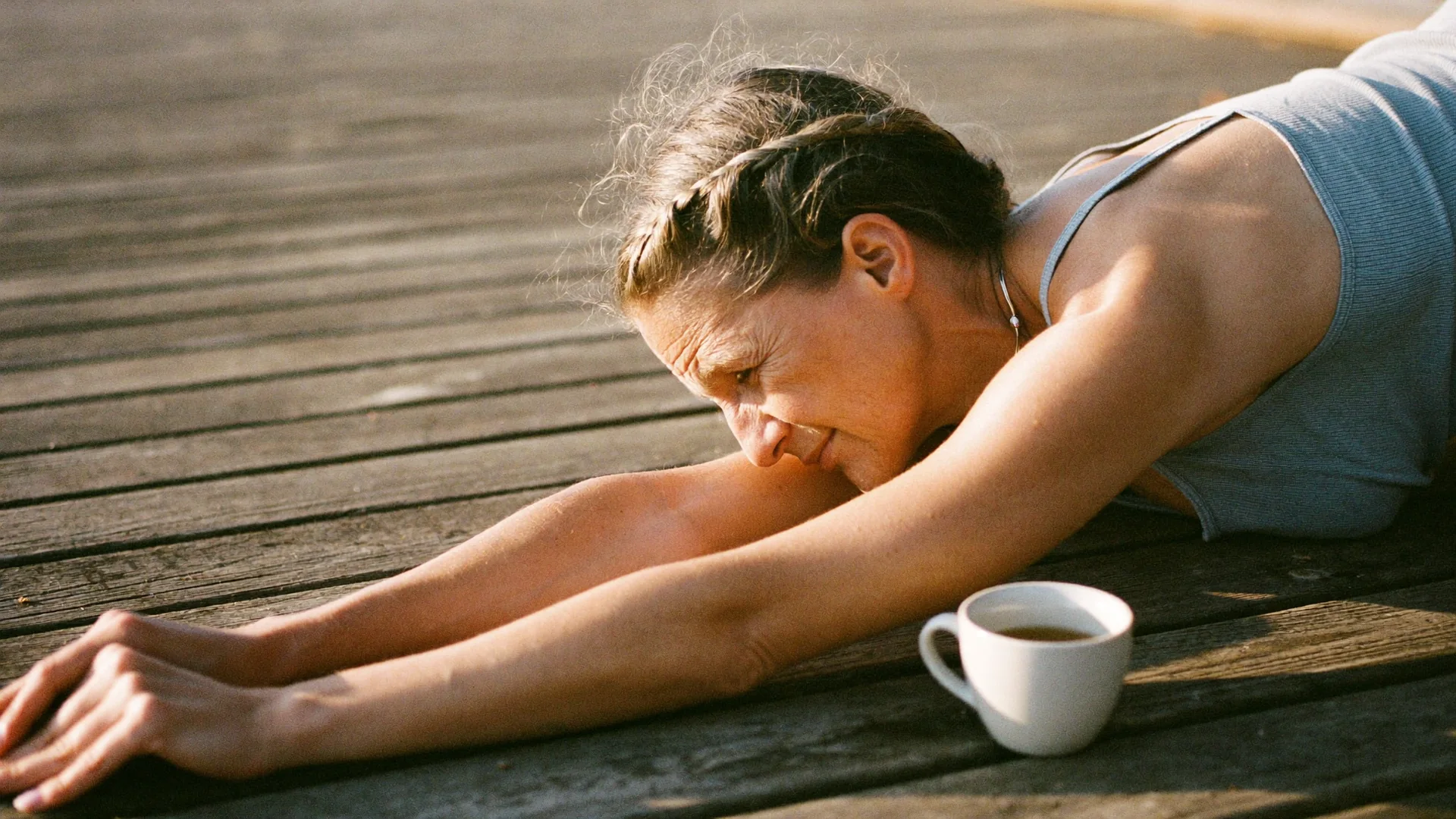 A woman in her forties stretching on a wooden deck at sunrise with a cup of tea beside her